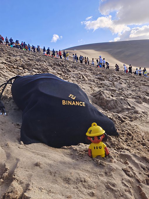 A photo taken at a sand dune showing a black Binance-branded bag and a Binance mascot figurine (yellow outfit with 'I ❤ U') in the foreground, with a line of people walking up the dune in the background.
