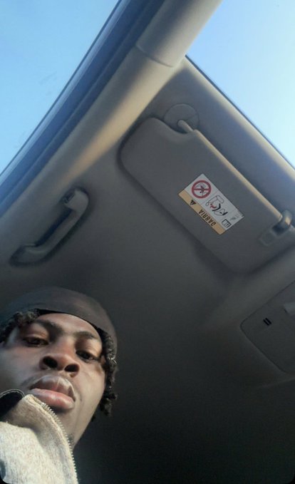 A low-angle selfie of a person inside a car, showing the sun visor with an airbag warning label and a view of the sky through the windshield.