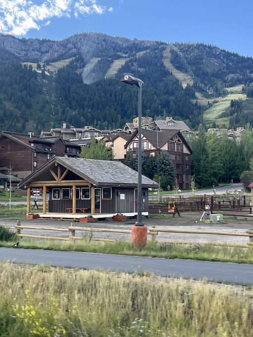 A scenic mountain village with rustic wooden buildings, chalets, and a ski lift visible on the forested mountainside in the background, under a partly cloudy sky.