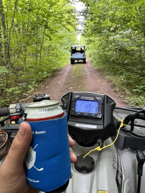 A first-person view from an ATV shows a hand holding a "Farmers" beer koozie while following another ATV down a wooded trail, with the digital gauge reading 30.0 — capturing the quintessential off-roading pastime that fits the "white boy activation" humor of suburban guys hitting the trails for some weekend fun.