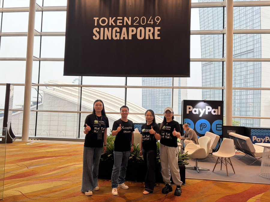 Four people wearing black t-shirts that read "Nại Tài Hai Yai" pose with thumbs up under a "TOKEN 2049 SINGAPORE" banner, with a PayPal display visible in the background.