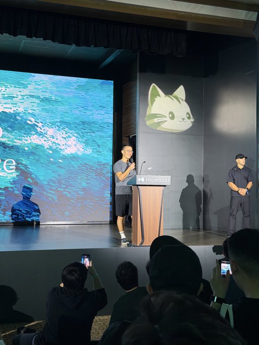 A man speaks at a podium branded with the Hyperliquid name, backed by a large screen displaying the project's signature cat logo, while audience members capture the presentation on their devices. The image aligns with the post's message, illustrating Hyperliquid's growing presence as a speaker addresses a packed room, emphasizing the "Dream bigger" sentiment.