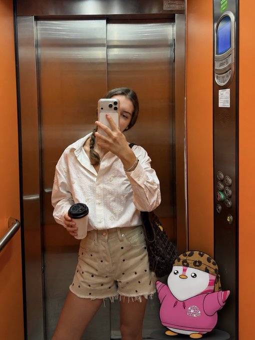 A mirror selfie of a woman in an elevator, wearing a white button-up shirt and beige polka-dot shorts, holding a coffee cup and phone, with a cartoon penguin sticker in the corner.