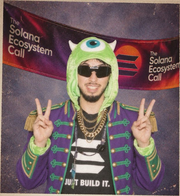 A man in a green Mike Wazowski (Monsters Inc.) hood and purple military-style jacket poses with peace signs in front of a "The Solana Ecosystem Call" banner.