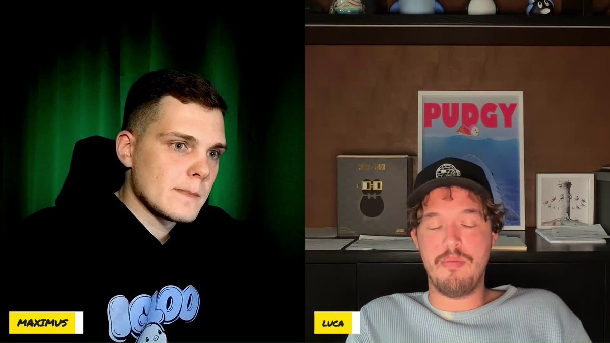The image shows two men sitting next to each other, with one man wearing a black hoodie and the other wearing a white shirt. The background features a poster for "Puggy" along with some books and a clock. There are also several books scattered around the room.
