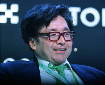 A man with glasses and dark hair, wearing a suit and a small lapel microphone, smiles while speaking at what appears to be a conference or panel event with blurred text visible in the background.