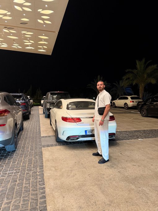 A man poses casually beside a sleek white luxury coupe parked under a decorative canopy at night, with palm trees and other vehicles visible in the background. The car displays a license plate ending in "NNE" as he greets his followers with a relaxed evening "Gm friends" vibe.