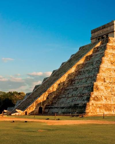 The ancient Mayan pyramid of El Castillo at Chichen Itza stands majestically against a blue sky, with warm sunlight illuminating its stone steps.