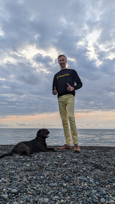 A man wearing a black "BINANCE" sweatshirt and yellow pants standing on a rocky beach giving two thumbs up, with a black dog lying beside him under a cloudy sunset sky.