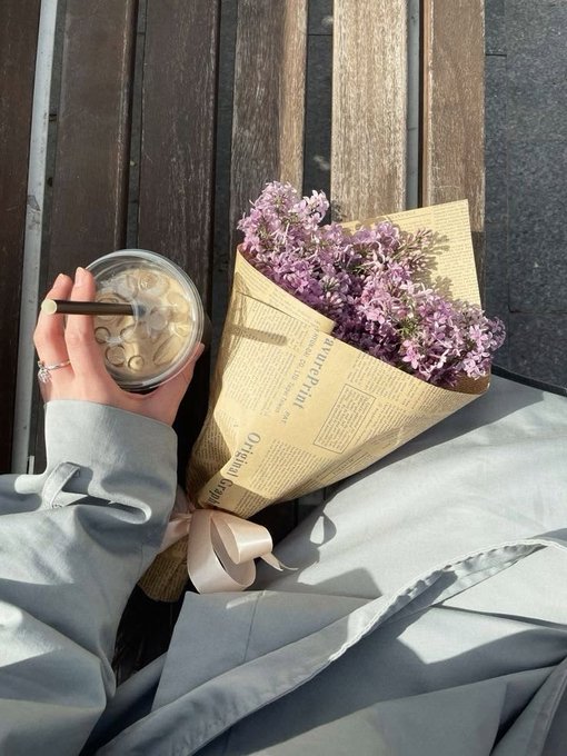 A person holding an iced coffee and a bouquet of purple flowers wrapped in brown newspaper-style paper, sitting against a wooden background.