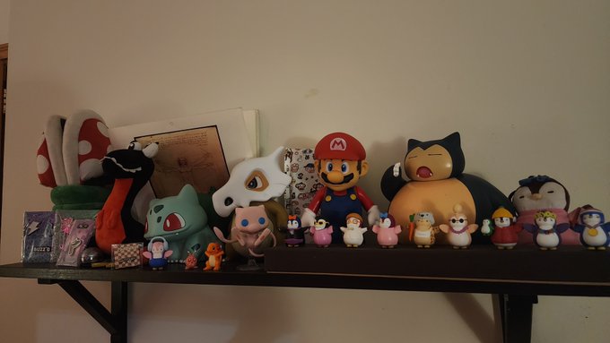 A shelf displaying a collection of plush toys including a Piranha Plant, Mario, Snorlax, Bulbasaur, and various small figurines in a softly lit room.