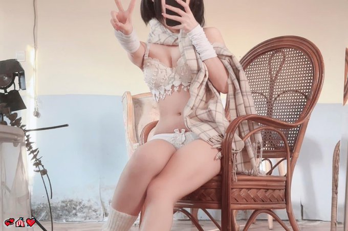 A woman in white lace lingerie and an open plaid shirt sits on a rattan chair, holding up a peace sign with one hand while taking a mirror selfie with her phone, with small pink heart graphics at the bottom.