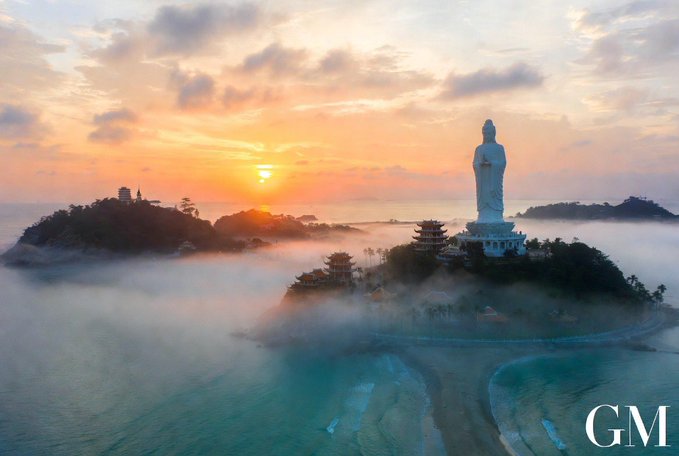 An aerial sunset view shows a large white Buddha statue standing on a misty coastal mountain above the sea, with 'GM' watermarked in the corner.