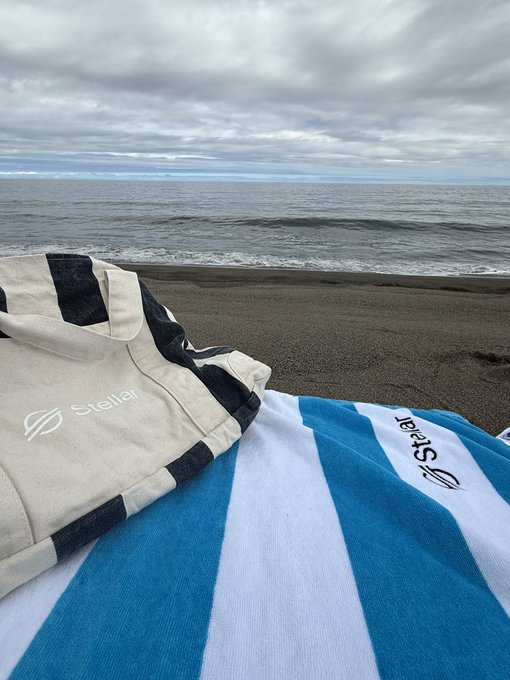 A view from a beach towel showing the ocean under a cloudy sky, with a black and white striped 'Stellar' bag and a blue and white 'Stellar' towel in the foreground.