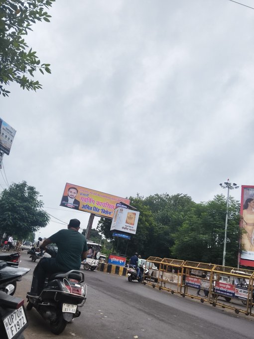 A street scene in India shows a man riding a motorcycle with the license plate 'UP70 F1972', passing under billboards including one for 'Amita Vidyalaya', with overcast skies above.