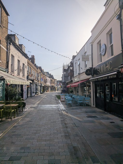 A street-level view of a paved European-style street lined with cafes and shops, including signs for 'GAIL's' and 'SUMAC,' with outdoor seating and string lights overhead.
