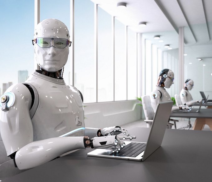 A humanoid robot wearing glasses types on a laptop in a modern office setting, with two other robots working at desks in the background.