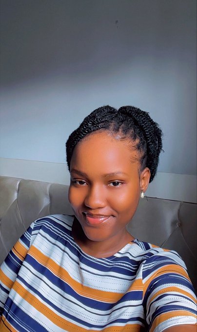 A selfie of a young woman with braided hair, wearing a striped orange, blue, and white shirt and small pearl earrings, smiling at the camera.