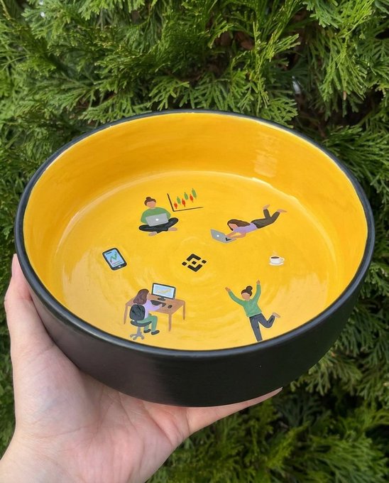 A hand holds up a yellow ceramic bowl decorated with illustrated figures working and relaxing around a central Binance logo, set against green foliage.