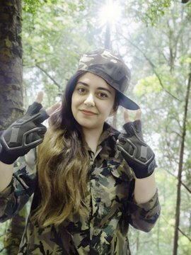 A young woman wearing a camouflage cap, matching jacket, and black tactical gloves makes peace signs with both hands in a forest setting.
