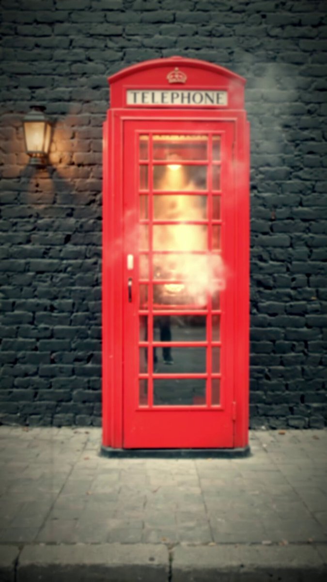 The image features a red telephone booth situated against a brick wall. The telephone booth has a lit interior that appears to be emitting smoke from its top. There are no people visible around the phone booth, suggesting an empty or deserted area. The surrounding environment includes a street lamp and a building with a brick facade. A caption is provided below the image, which reads: "Is this a clue for RED = Miami? Last month at Consensus in Miami, Florida we had a BAYC community event (presented by @apecoin & @0xBoringYachts) at a speakeasy located behind a RED phone booth."