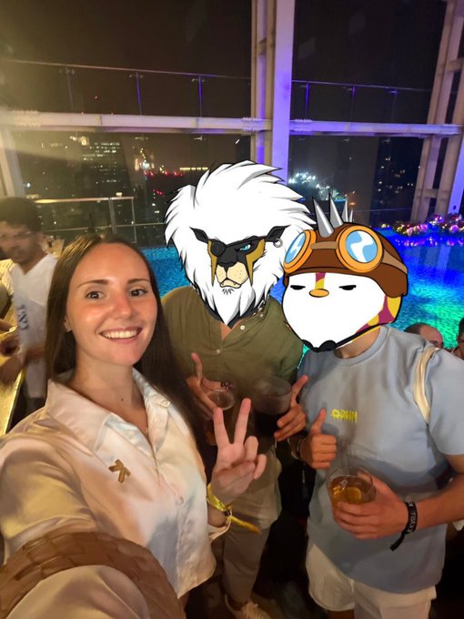 A woman smiles for a selfie with two friends at an evening rooftop gathering, all flashing peace signs and holding drinks. Two cartoon character stickers (a white lion and a creature with goggles) have been placed over the faces of the people in the background, matching the playful spirit of the "Gm and happy Friday! Look who I met yesterday" caption.