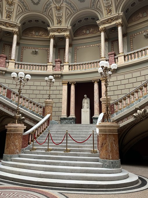 Grand interior of an ornate building featuring an elegant marble staircase with curved steps, a white statue at the landing, decorative balustrades, classical columns, and vintage street lamp-style light fixtures.