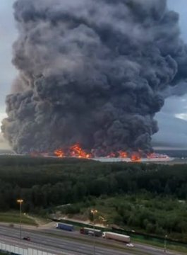 A photograph of a large industrial fire producing massive dark smoke plumes rising into a cloudy sky, with highways and trees visible in the foreground.