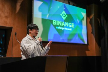 A man in glasses and a white jacket speaks at a podium with a microphone, presenting in front of a large screen displaying the Binance logo and 'exchange the world' tagline.