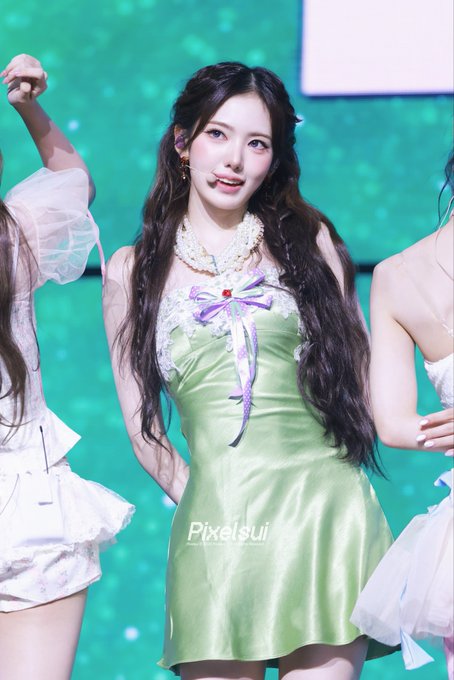 A performer with long dark wavy hair wears a green dress with pearl detailing and a purple ribbon, smiling on a teal-lit stage. Watermark reads 'Pixelsui'.