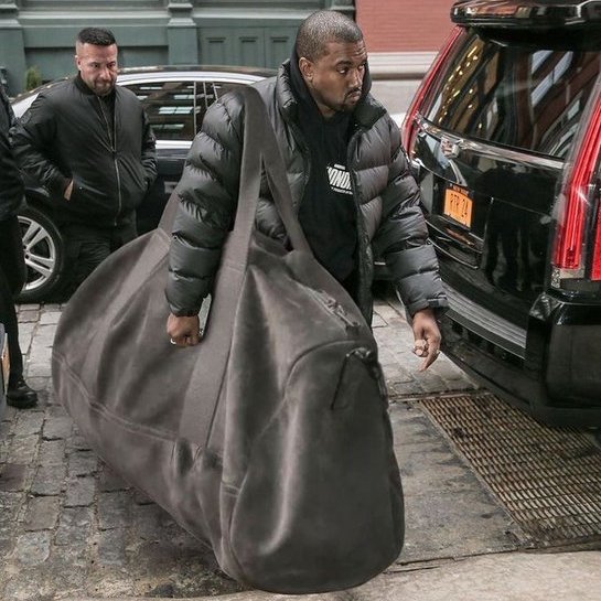 The image shows a man in a black puffer jacket struggling to carry an oversized gray duffel bag, standing on a cobblestone street next to a black SUV with a visible New York license plate—visually representing the concept of a "biggest bag" referenced in the post about memecoin holdings.