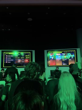 Photo taken from the audience at an event showing two large screens displaying digital content, with attendees seated in a dimly lit auditorium.