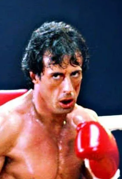 A shirtless Sylvester Stallone as Rocky Balboa, wearing red boxing gloves and showing a cut above his eye, looking battered but determined in the boxing ring. The image visually reinforces the post's metaphor comparing Bitcoin to the resilient boxer who repeatedly overcomes setbacks, embodying the asset's history of bouncing back from six major 50% drawdowns.
