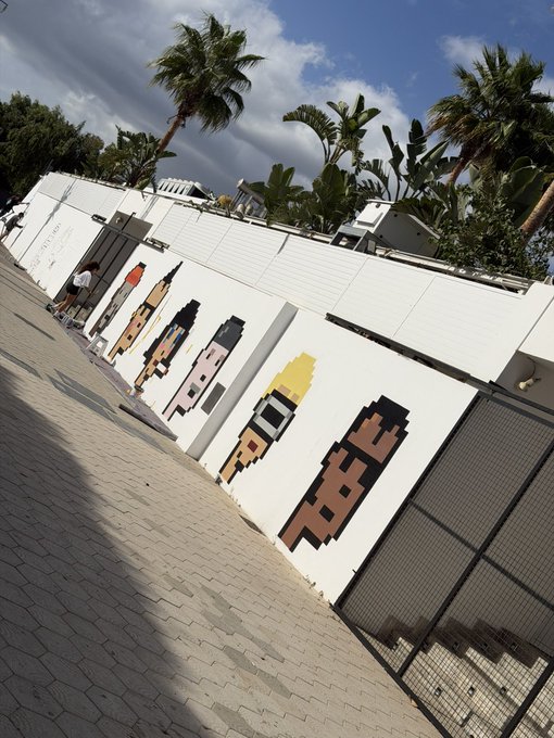 An outdoor scene showing a white wall with pixel art designs alongside palm trees and tropical plants under a cloudy sky.
