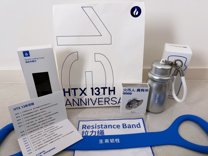 A collection of HTX 13th anniversary merchandise and promotional items laid out on a surface, including a resistance band, a water bottle, a sports cooling towel, and packaging boxes with "HTX 13TH ANNIVERSARY" text.