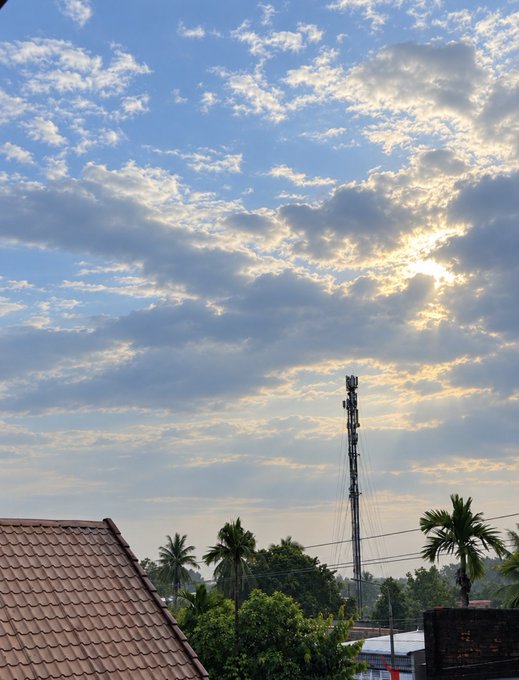 A photograph of a vast sky filled with scattered clouds with the sun shining through, above a communication tower, palm trees, and the tiled roof of a building in the foreground.