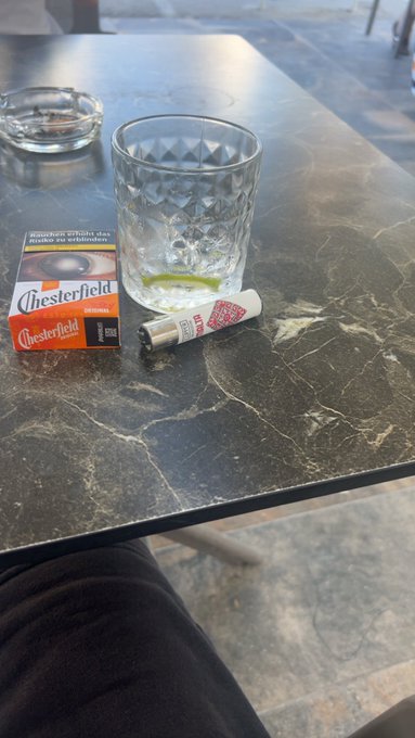 The image features a table with various items, including an ashtray containing a cigarette, a glass of water, and some Chesterfield cigarettes. The scene also includes a chair positioned near the table. The text "life is good my friends $btc short is printing always remember to pay yourselfs" is present in the image as well."