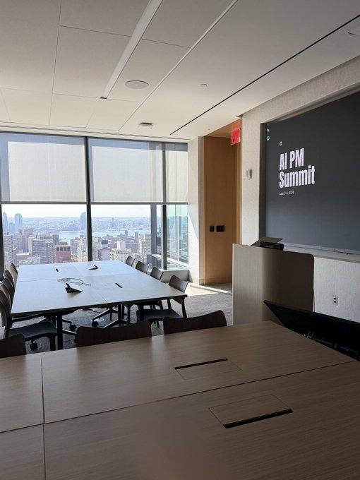 The image shows an office setting with a large screen TV mounted on the wall displaying "AI PM Summit" written in white letters. The room features several tables and chairs arranged around the space, suggesting a collaborative work environment. There are also multiple TVs visible within the room, indicating that this is likely a shared workspace or conference area for various projects.