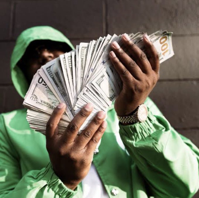 A person wearing a green hooded jacket holding a large fan of US dollar bills in front of their face, wearing a gold and silver watch.
