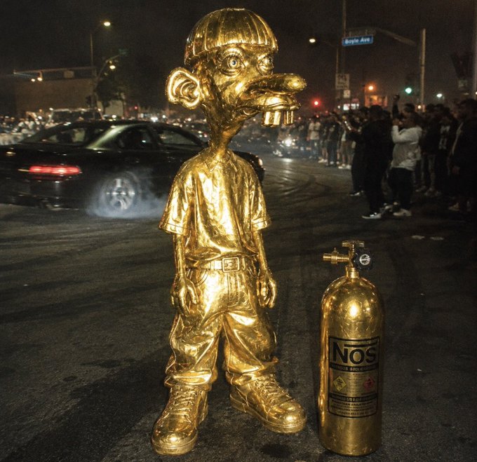 A golden statue of a cartoonish boy stands next to a gold-painted nitrous oxide tank labeled 'NOS', set against a nighttime street scene with a crowd and cars.