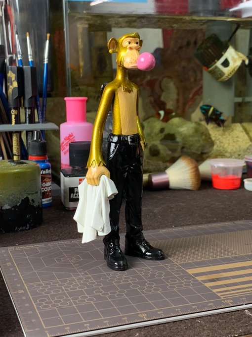 A custom-painted figurine of a giraffe-headed figure in a gold shirt and black pants, blowing a pink bubble gum bubble, standing on a cutting mat among art supplies.