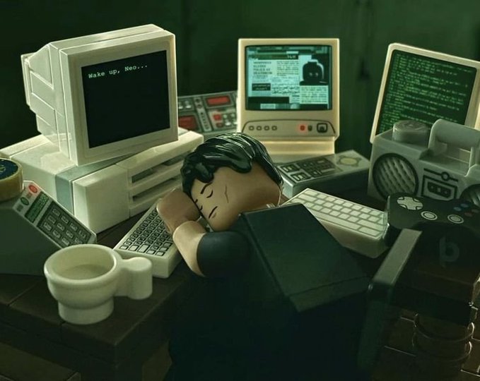 A LEGO-style diorama showing a minifigure sleeping at a desk surrounded by vintage computer monitors, one displaying "Wake up, Neo..." and another showing green code.