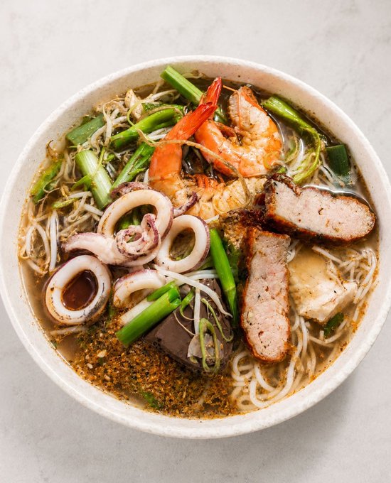 A top-down photo of a bowl of noodle soup containing shrimp, squid rings, pork slices, tofu, blood cubes, green onions, and bean sprouts in a clear broth.