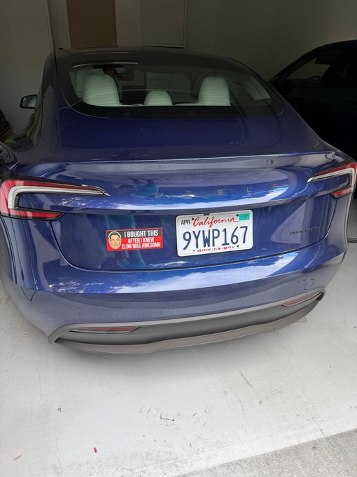 Rear view of a blue Tesla vehicle with a California license plate reading '9YWP167' and a bumper sticker stating 'I BOUGHT THIS AFTER I KNEW ELON WAS AWESOME'.