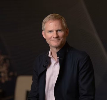 The image shows a portrait of a man with short gray hair, wearing a dark blazer over a pink button-down shirt, smiling slightly at the camera. Based on the accompanying post identifying him as BitGo CEO Mike Belshe, the portrait accompanies his statement urging lawmakers to support the CLARITY Act to prevent future exchange collapses like FTX.