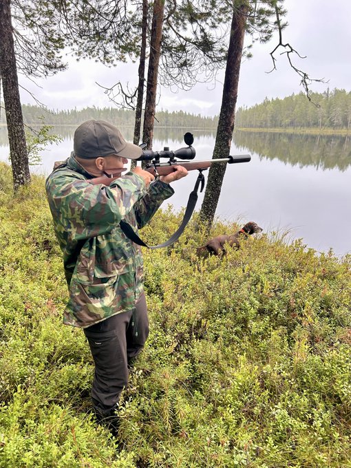 A hunter in camouflage aims a scoped rifle while a dog prowls through the brush beside a tranquil lake in a forested area. The image's rural hunting trip atmosphere matches the post's witty jab about abandoning the chaotic financial markets for the quiet outdoors.