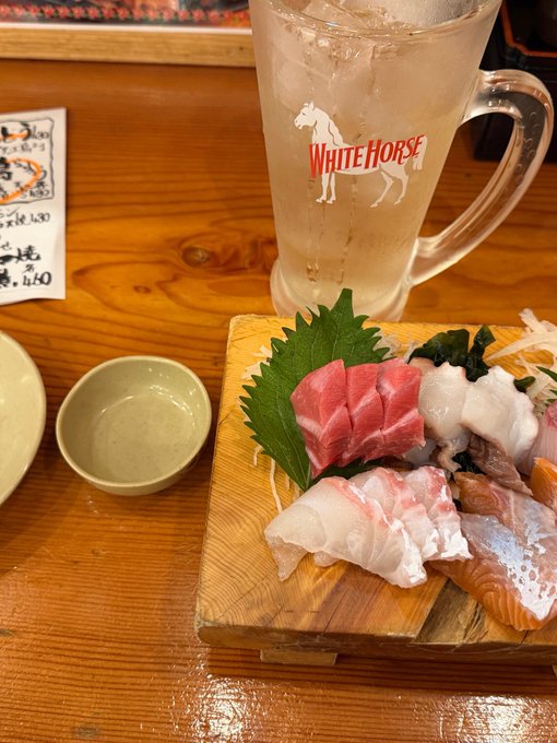 The image features a wooden dining table with various food items, including sushi, sashimi, and a glass of beer. The sushi has been cut into pieces and arranged on a wooden platter or cutting board. A bowl can also be seen in the scene. There is a cup next to the beer mug, which appears to contain ice-cold water. The overall setting suggests that this could be a casual dining experience at a restaurant or a gathering with friends where people are enjoying sushi and drinks together.
