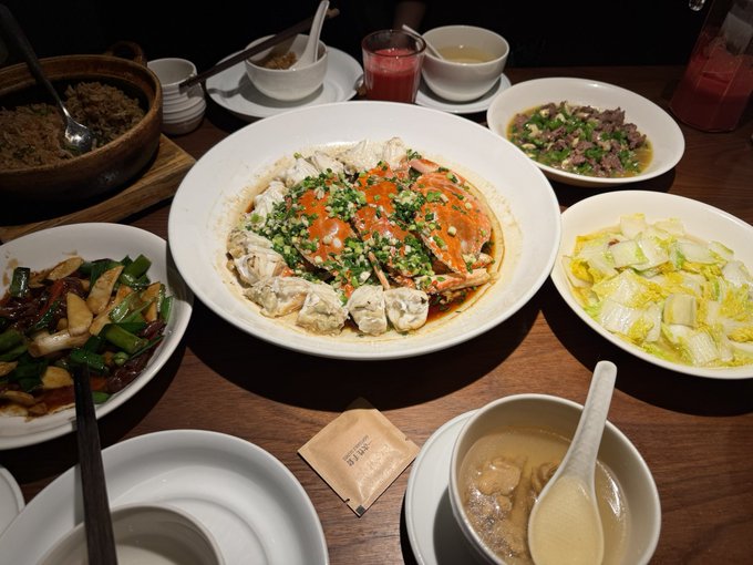 A spread of Chinese dishes on a wooden table, featuring a central plate of steamed crab garnished with garlic and scallions, surrounded by stir-fried greens, meat with scallions, baby bok choy, rice, and soup.