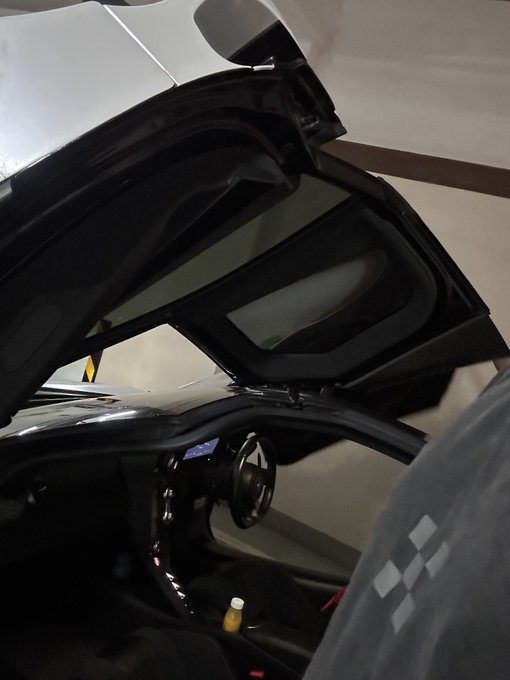The image shows a car with its hood open, revealing the interior of the vehicle. The car appears to be black and white or silver, depending on the lighting conditions. A person can be seen inside the car, likely driving or preparing for a journey. There is also an image of a person's hand holding a cup in the background, possibly indicating that they are enjoying a beverage during their drive.