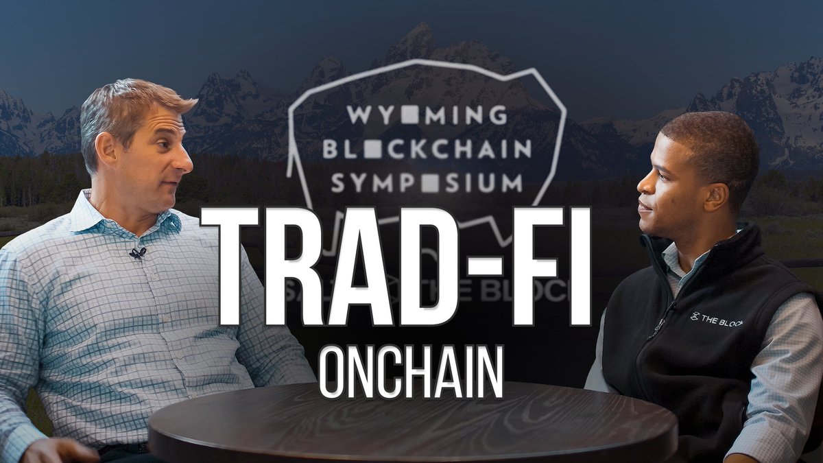 Two men seated at a table for an interview setting, with 'WYOMING BLOCKCHAIN SYMPOSIUM' and 'TRAD-FI ONCHAIN' overlaid on the image, with mountains in the background.